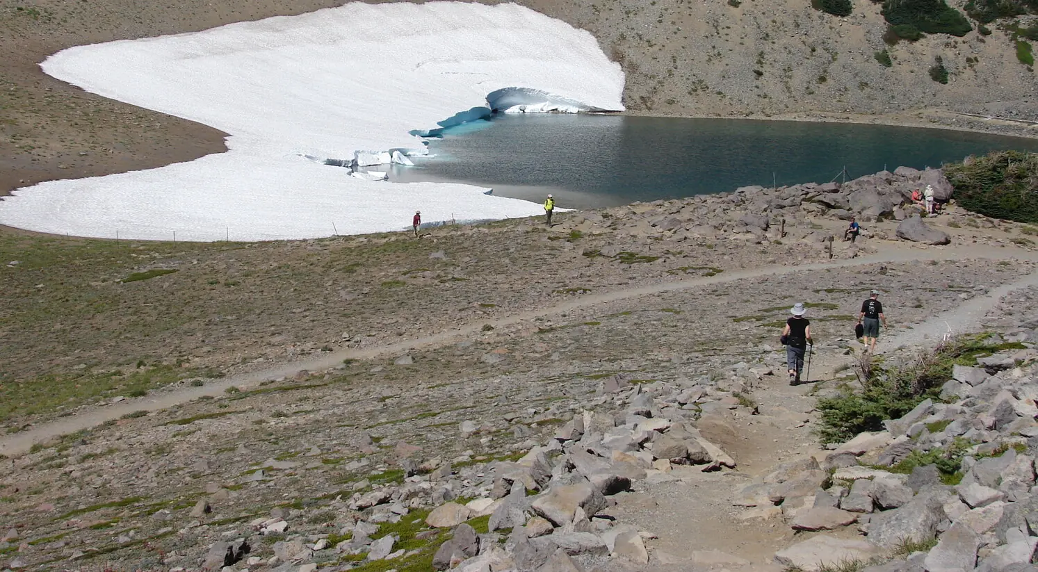 Hikers in summer clothing walking a gravel trail across open tundra above a small alpine lake, half of which is still covered by a thick collapsing snowfield