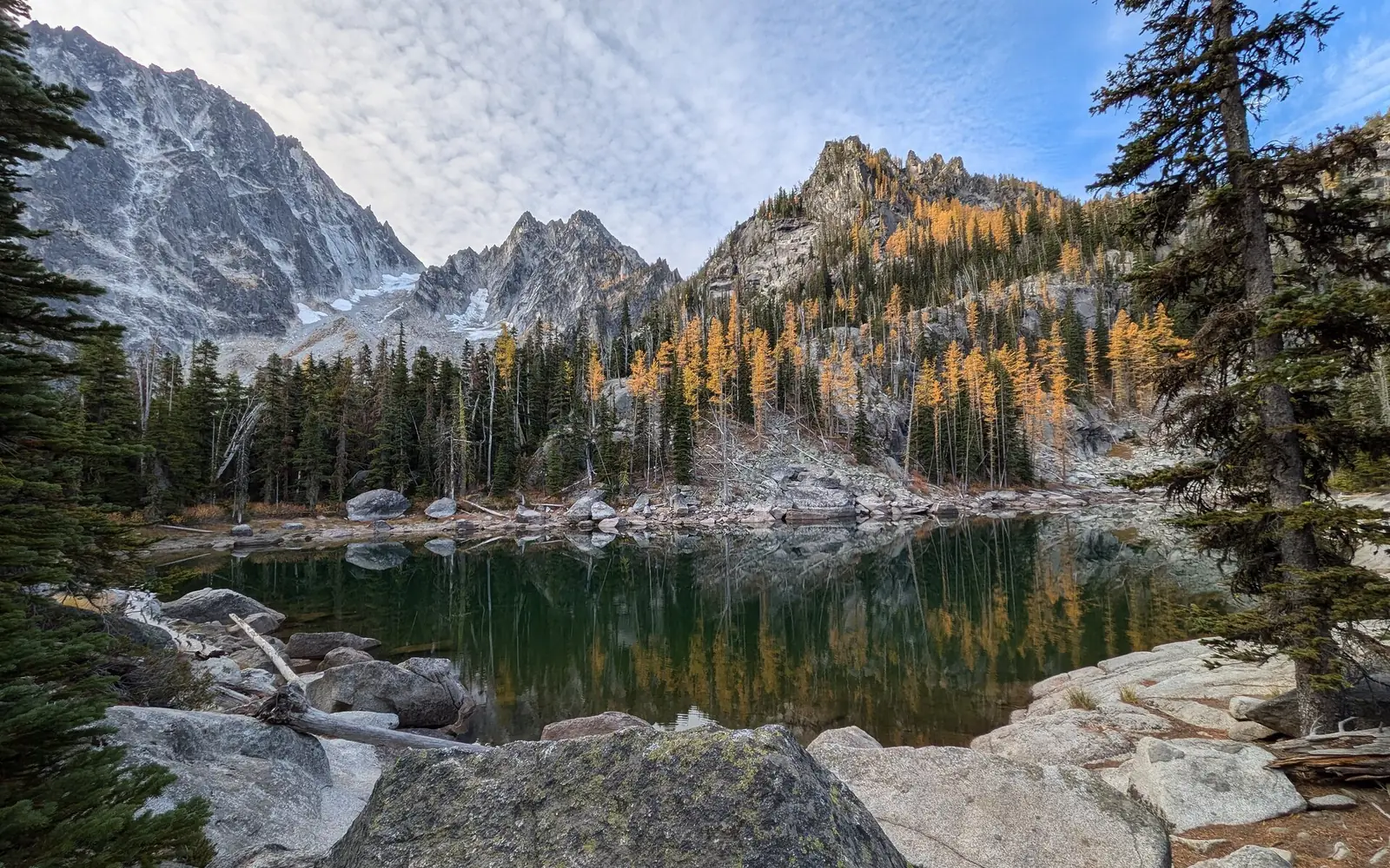 A still alpine lake reflecting a slope of golden larches, with jagged gray granite peaks and a patch of snow above the far shore