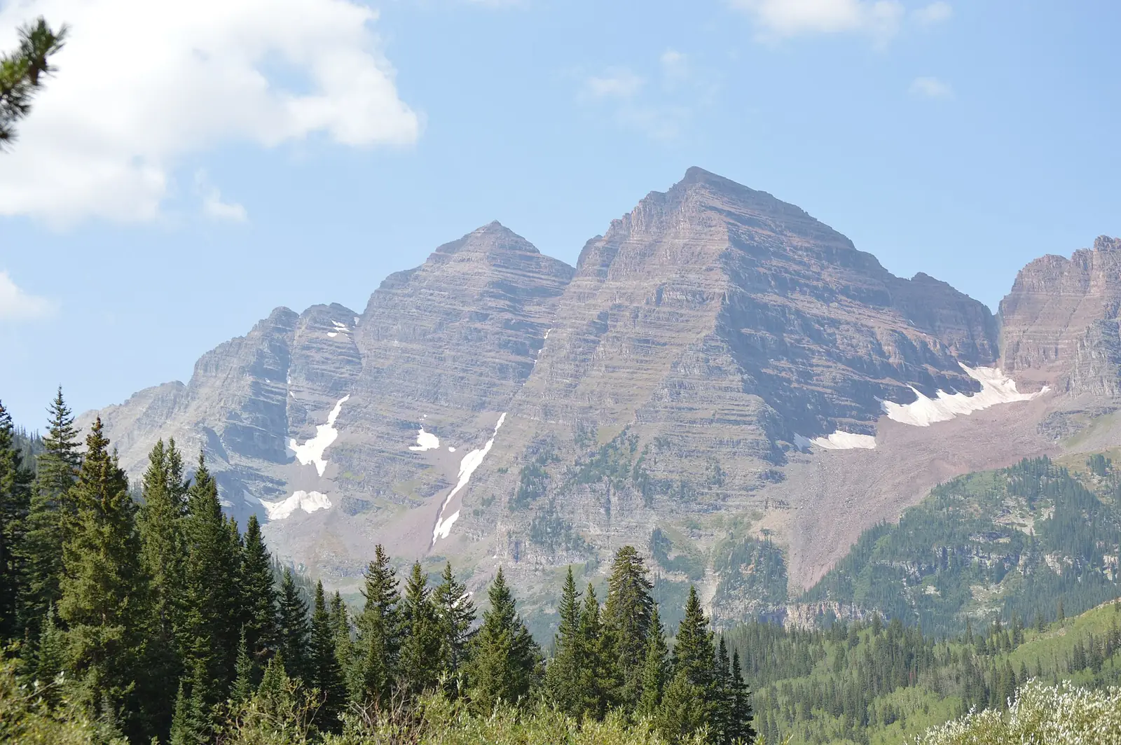 The twin maroon-colored summits of the Maroon Bells above dark spruce forest, streaked with old snow under a bright summer sky