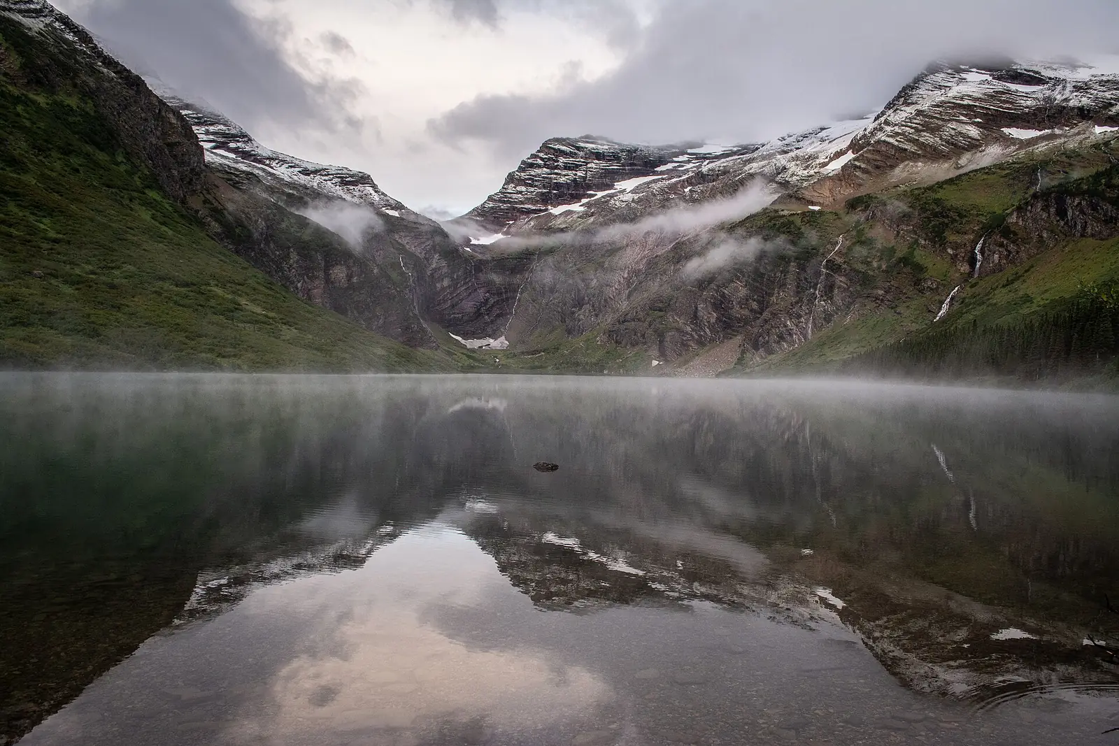 A still alpine lake holding a perfect reflection of a snow-streaked headwall, with mist lying on the water under low gray cloud
