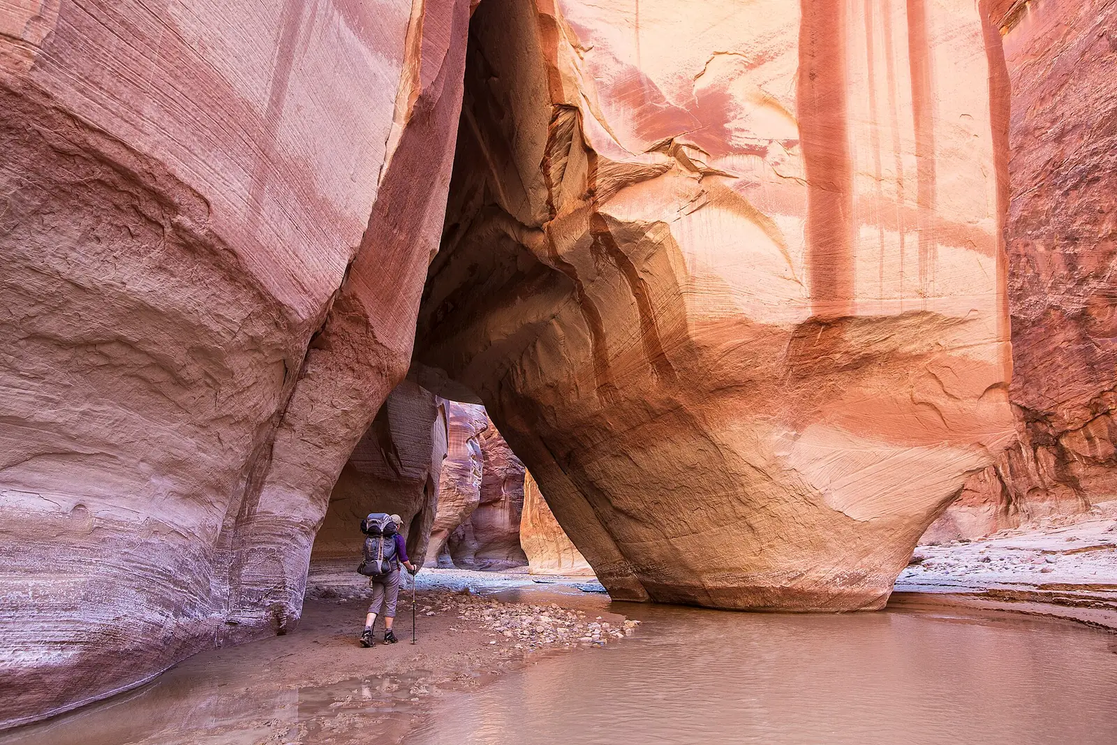 A backpacker standing on the sandy floor of a deep sandstone canyon, dwarfed by curving orange and pink walls that lean together overhead