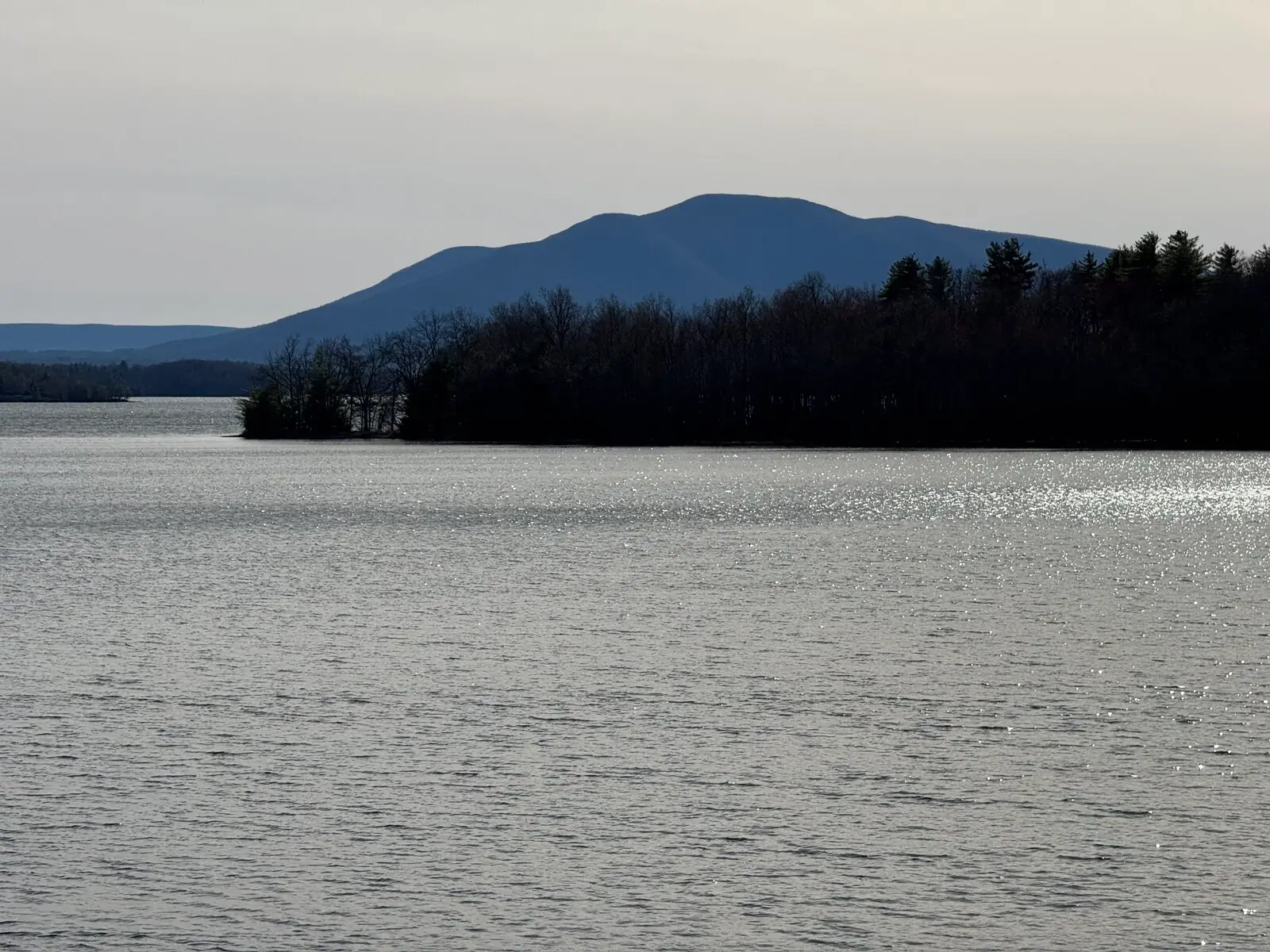 The long forested ridge of Ashokan High Point silhouetted beyond the flat silver water of the Ashokan Reservoir on an overcast day