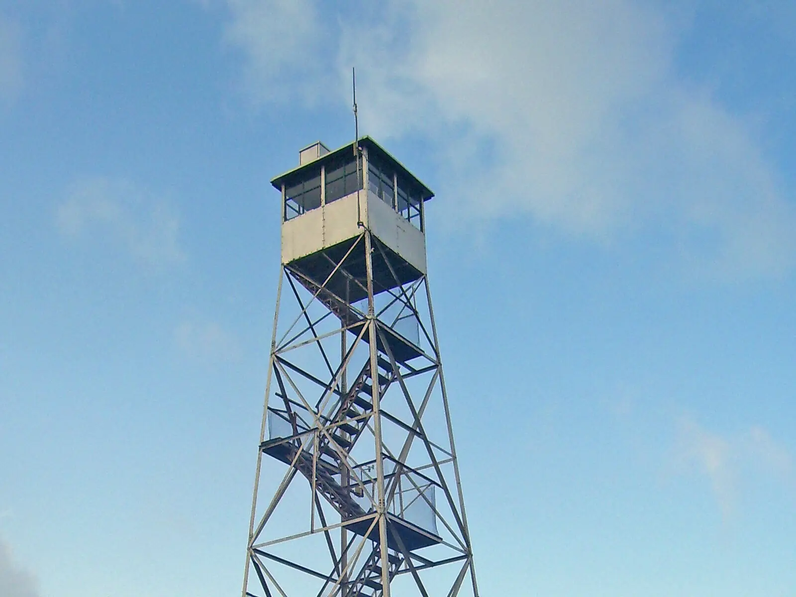 A restored steel fire tower with an enclosed glass cab at the top, rising above spruce trees against a blue sky with light cloud