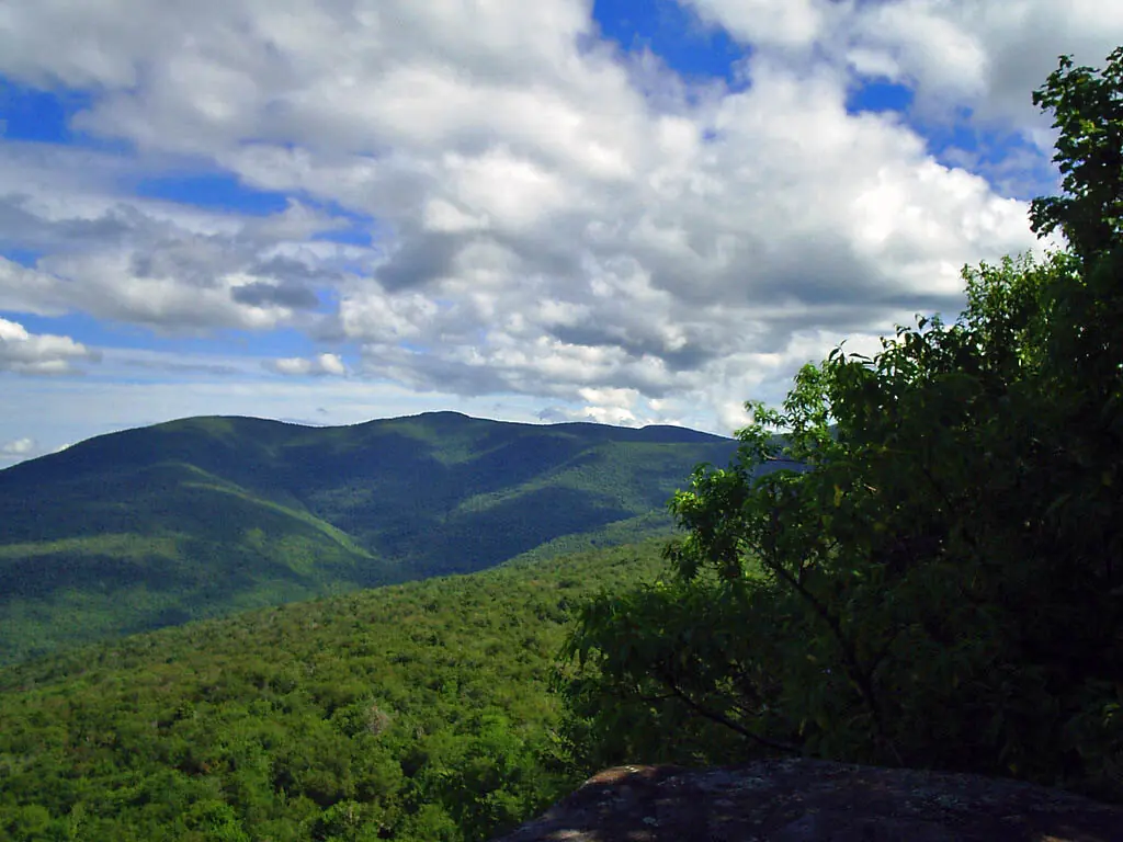 A wide summer view from a rock ledge over deep green forested valleys to the long ridge of Wittenberg and Cornell mountains under a cloudy sky