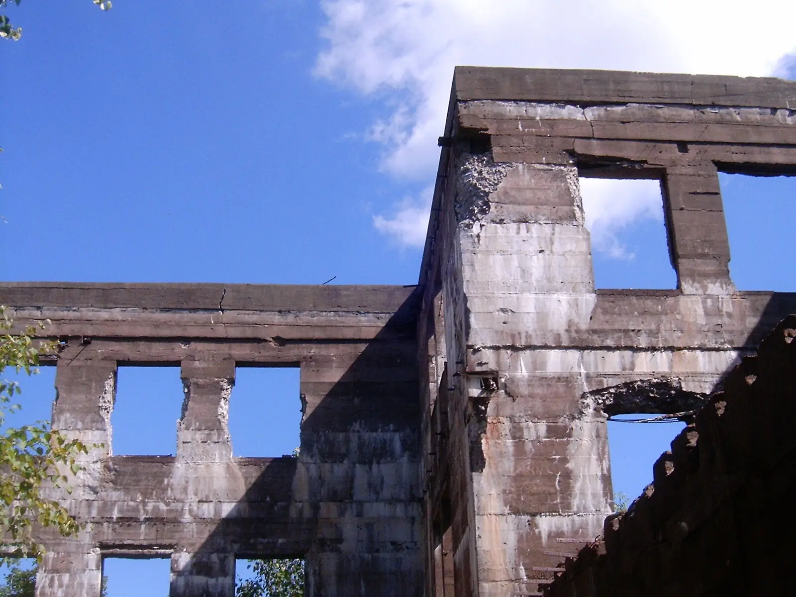 Roofless concrete walls of an abandoned hotel standing in summer woodland, empty window openings framing blue sky and trees