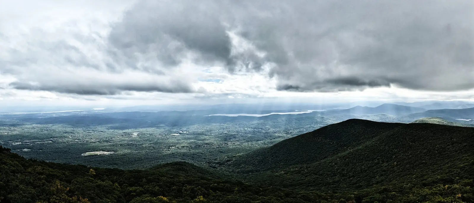 A sweeping view from the Overlook Mountain fire tower over the Hudson Valley, with shafts of light falling through heavy gray cloud onto the Ashokan Reservoir and forested ridges