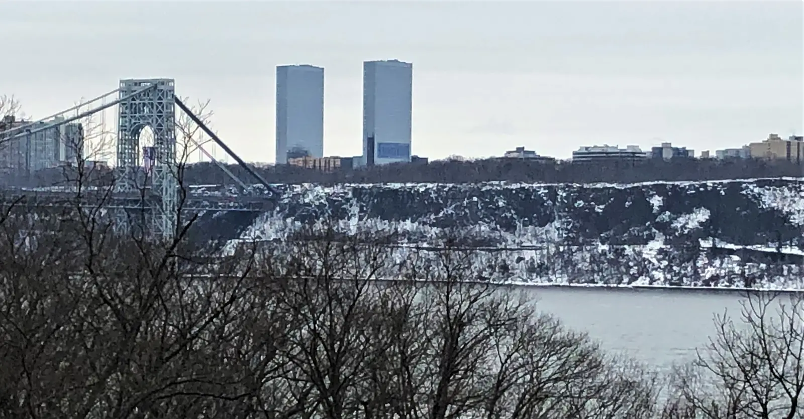 The New Jersey tower of the George Washington Bridge on a gray winter day, with the towers of Fort Lee behind it and the snow-streaked rock face of the Palisades running away to the right above the Hudson