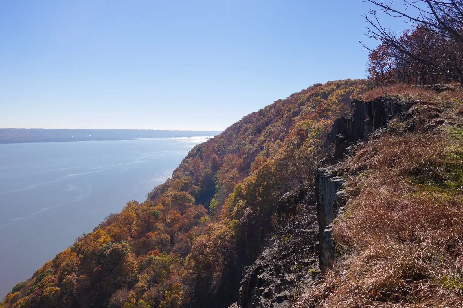 A basalt cliff edge on Hook Mountain in November, the wooded slope below it in orange and gold, the Hudson River wide and pale beyond and a bridge crossing it in the far distance