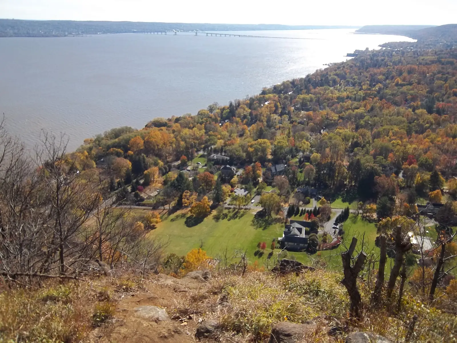 The view down from the top of Hook Mountain over the wooded village of Upper Nyack in full autumn color, the wide Hudson River beyond and a long bridge crossing it in the distance