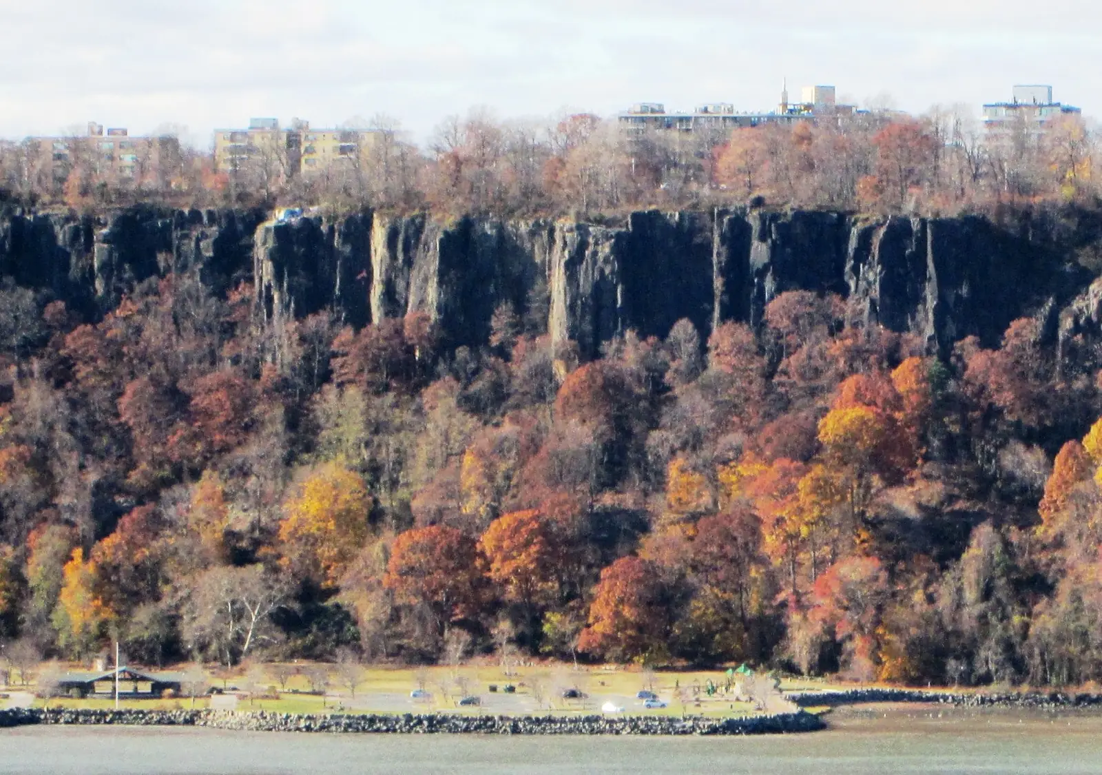 The wall of the Hudson Palisades seen across the river from Washington Heights in Manhattan, gray columnar cliffs above a slope of rust and orange November woods, with apartment buildings along the clifftop