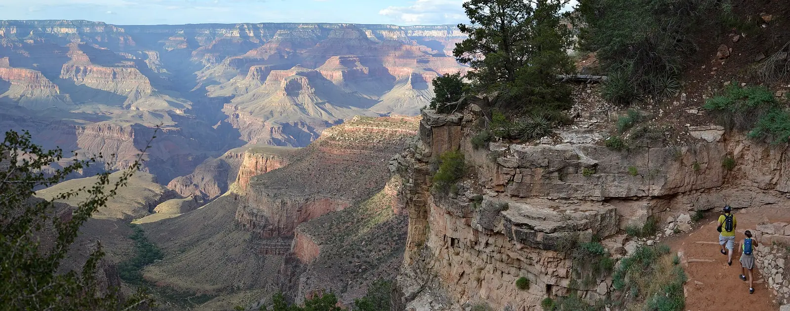Two hikers with daypacks walking up the wide upper section of the Bright Angel Trail along the base of a cliff, with the Grand Canyon opening out across the left of the frame