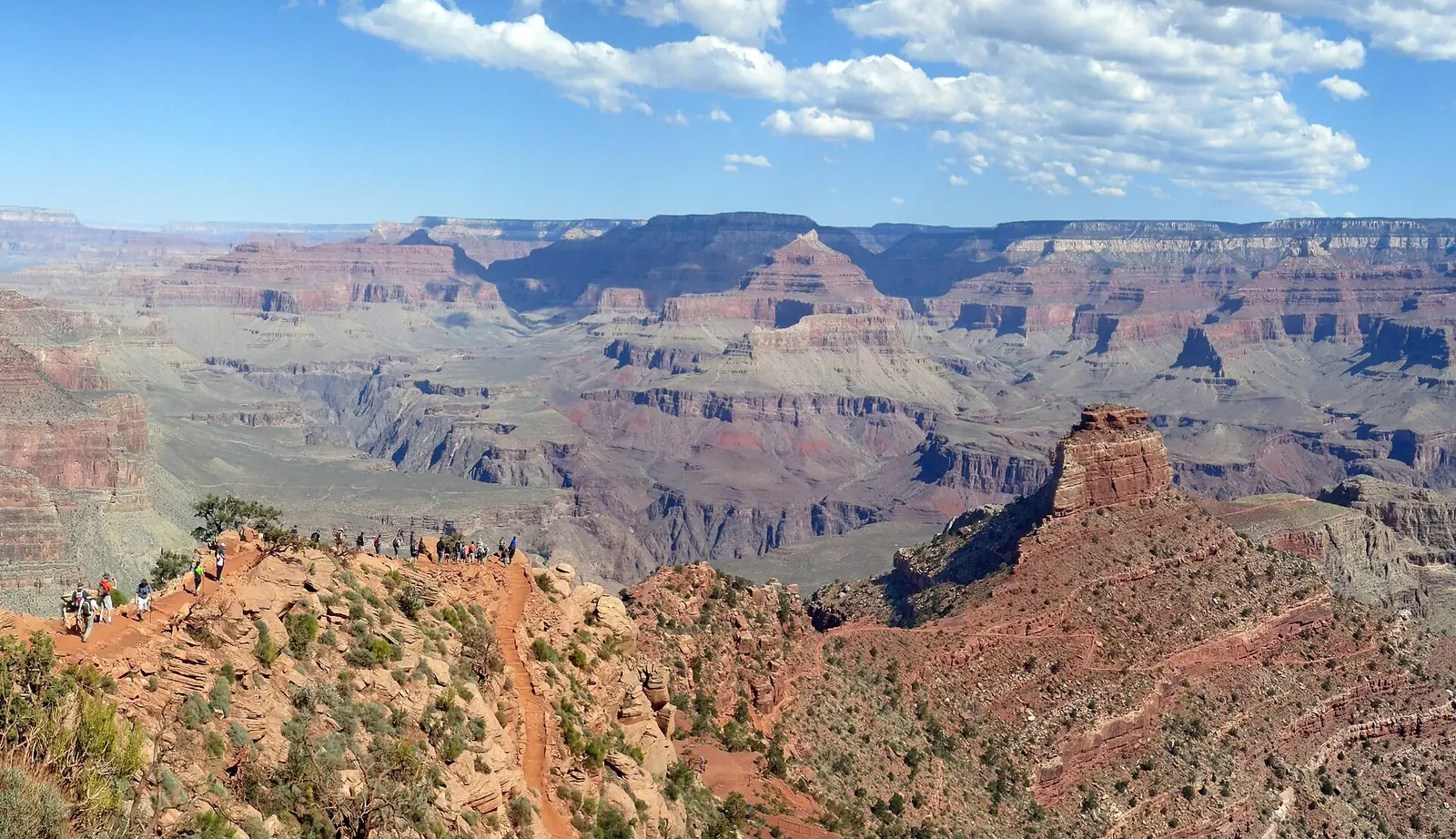 A line of hikers strung along the open red ridge of the South Kaibab Trail at Cedar Ridge, with a flat-topped butte to the right and the canyon stacked in layers behind