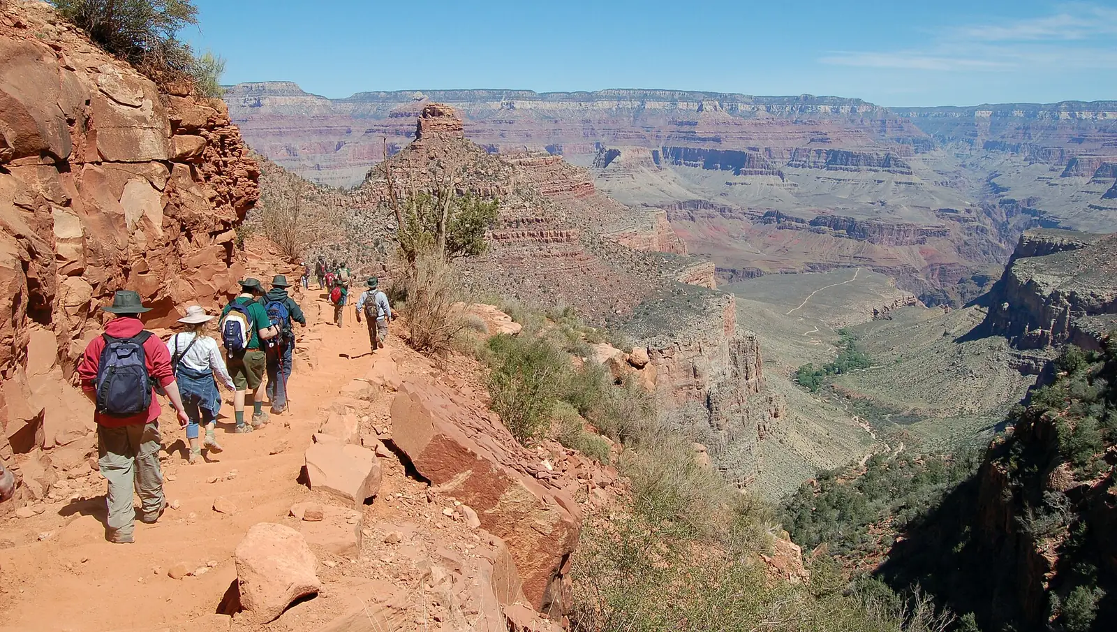 A line of hikers with daypacks walking down a red dirt trail cut into a cliff below the South Rim of the Grand Canyon, with layered canyon walls stretching to the horizon