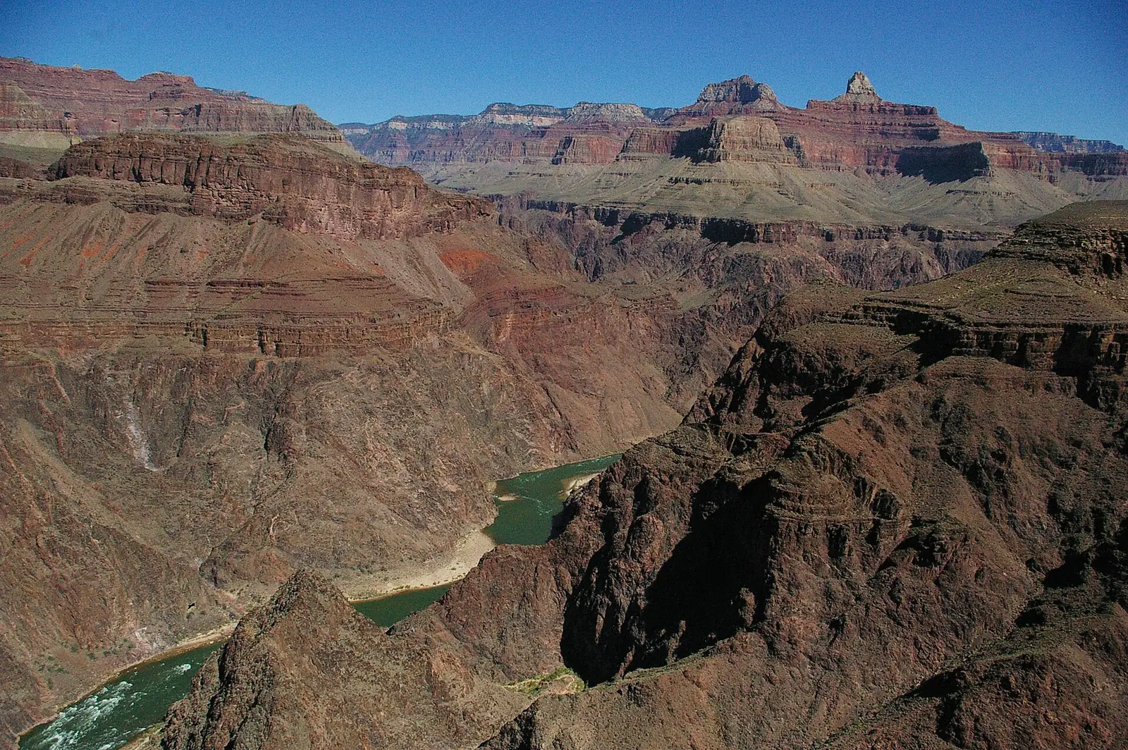 The green Colorado River running through the dark rock of the inner gorge of the Grand Canyon, seen from above, with red and gray layered cliffs and buttes rising beyond