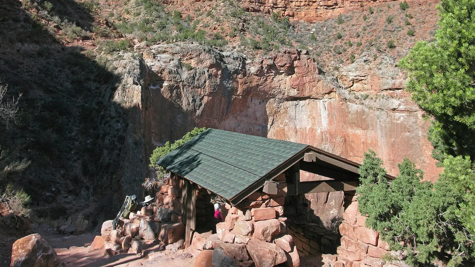The stone 3-Mile Resthouse with a green shingled roof standing on a bench of the Bright Angel Trail, with hikers and packs beside it and a red cliff wall rising behind