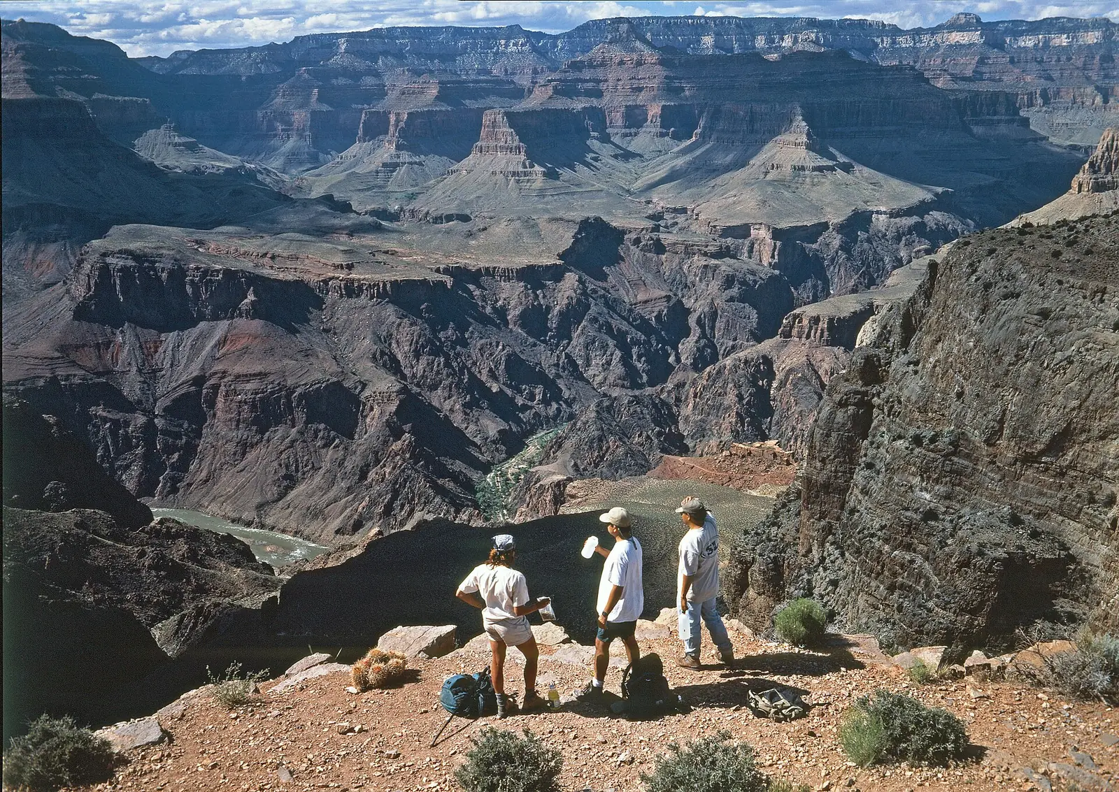 Three hikers standing at a rocky viewpoint on the South Kaibab Trail looking down into the dark inner gorge, with a green stretch of the Colorado River visible far below