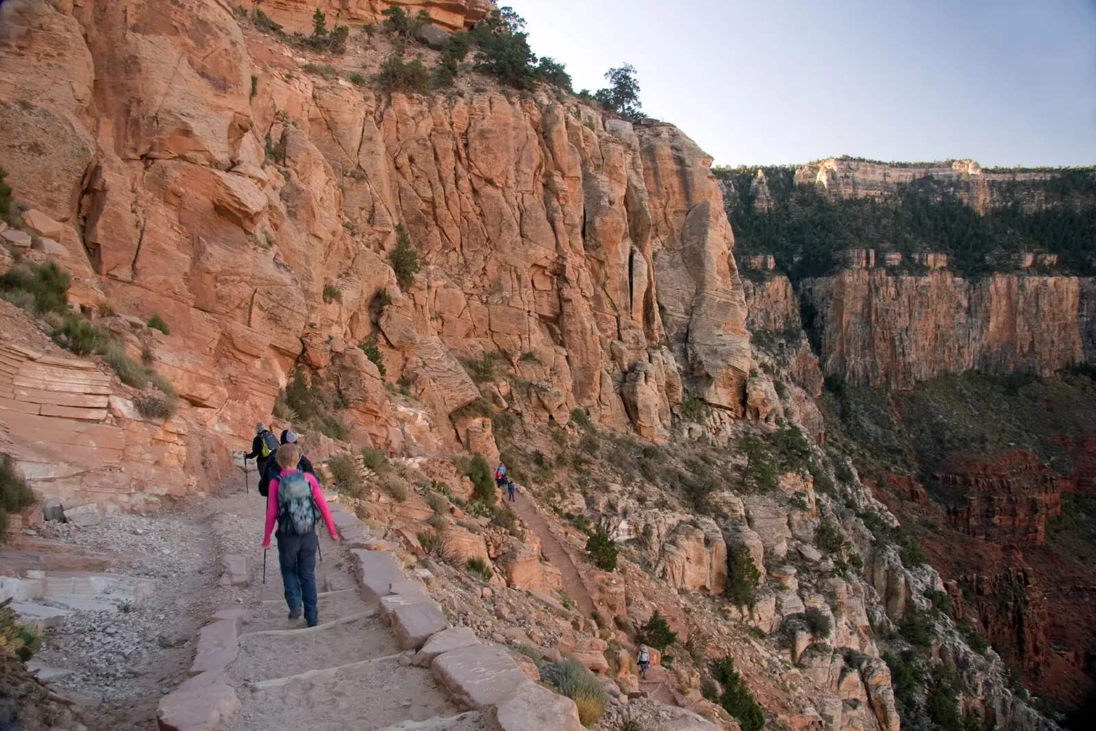Hikers with trekking poles descending a stone-cut section of trail below the South Rim of the Grand Canyon, with a huge orange cliff face beside them