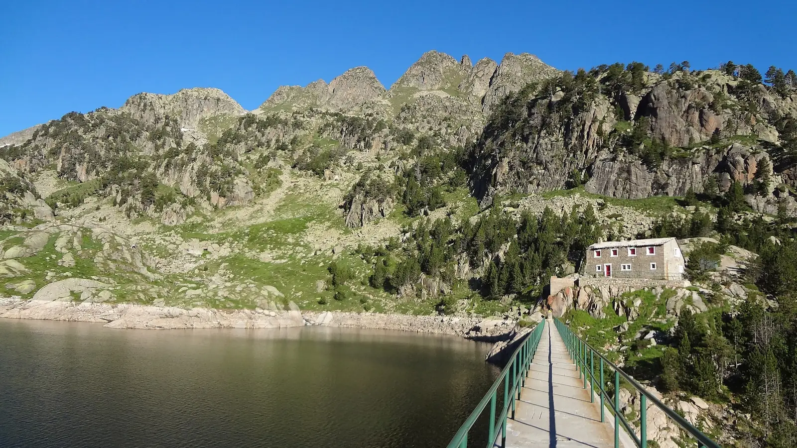 A narrow footbridge along a dam wall at Estanh Major de Colomèrs in the Catalan Pyrenees, leading to a small stone refuge with red shutters on a rock outcrop, with pine forest and granite ridges beyond