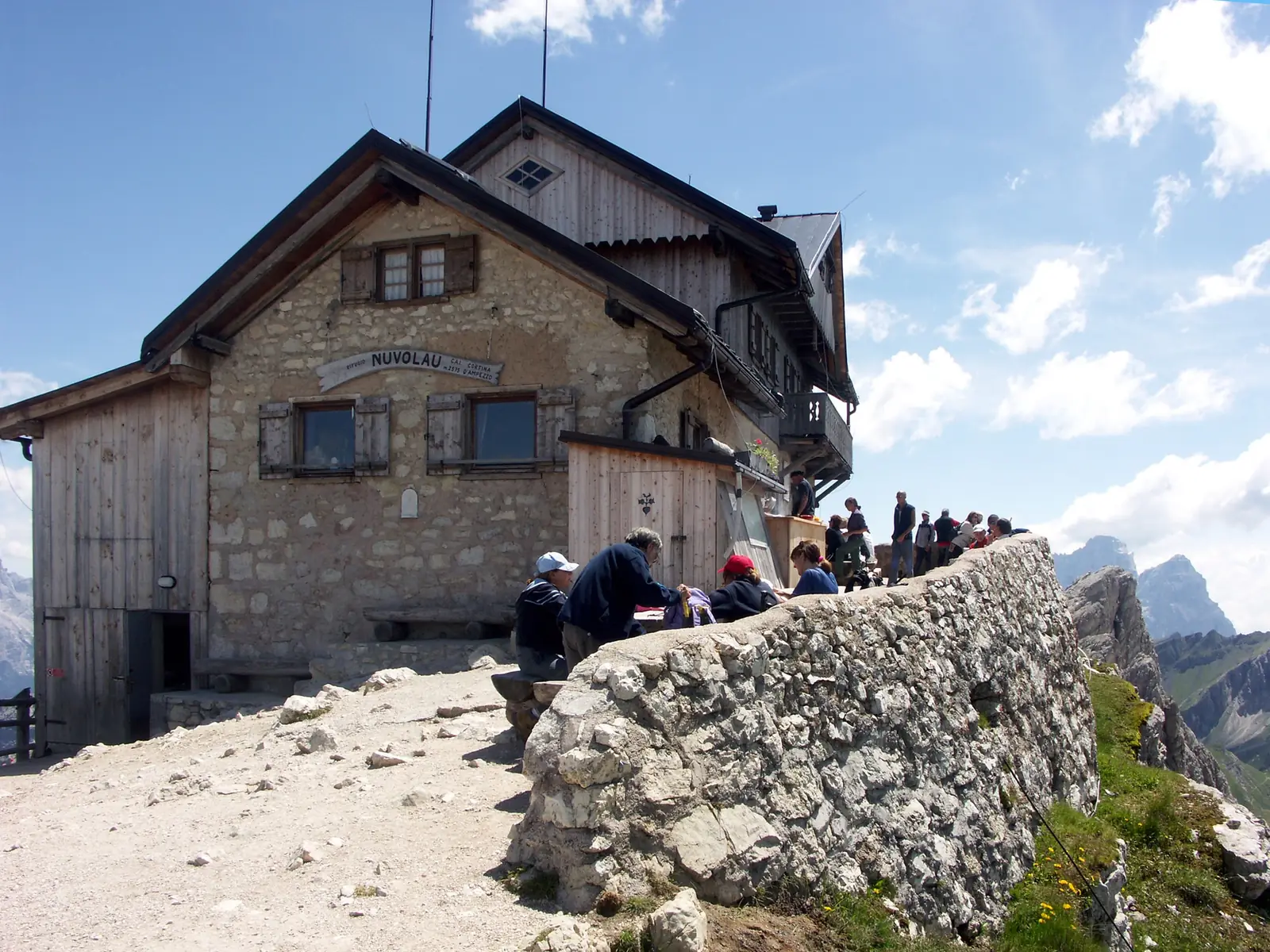 The Rifugio Nuvolau, a stone and timber hut with a painted nameplate on its gable, standing on a bare rocky summit in the Dolomites, walkers sitting at benches beside a low stone wall along the edge and pale limestone peaks in the distance