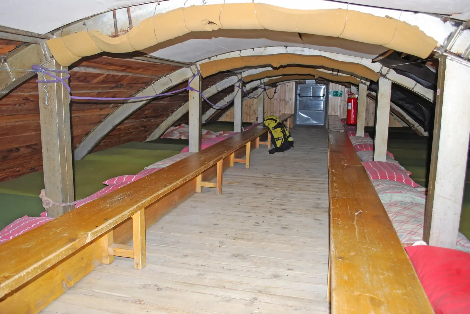 A long attic dormitory in an Italian mountain refuge, with two rows of mattresses, pillows and folded blankets under a curved wooden roof, a bench down the middle and a rucksack hanging from a post
