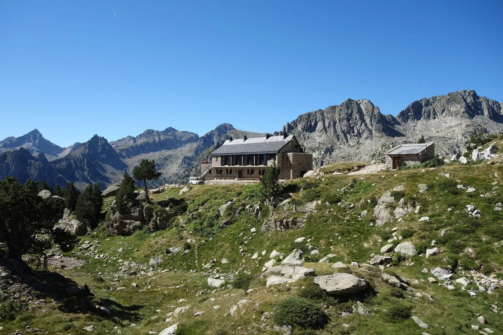 The stone Refugi d'Amitges standing on a grassy shoulder in the Catalan Pyrenees, with jagged granite spires and scree slopes rising behind it under a clear blue sky