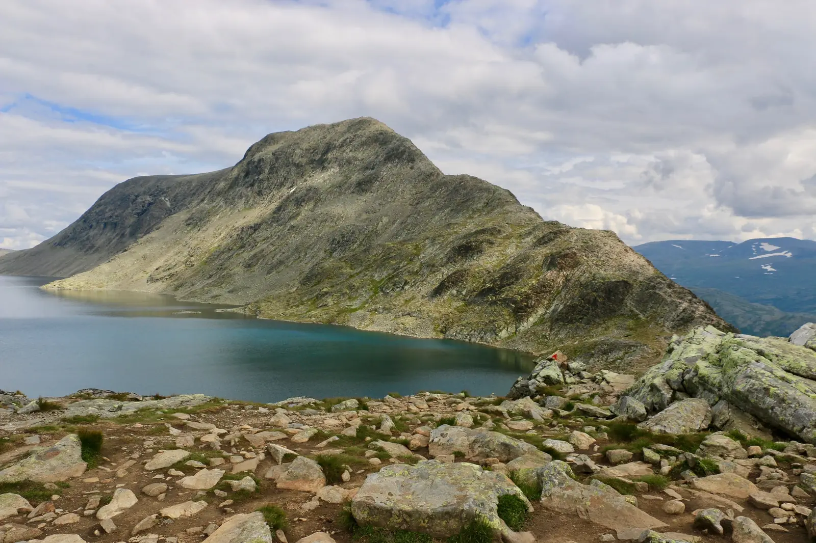 The Besseggen ridge in Jotunheimen falling away to the deep blue-green water of Lake Gjende, with a red painted waymark on a boulder in the foreground and broken cloud above