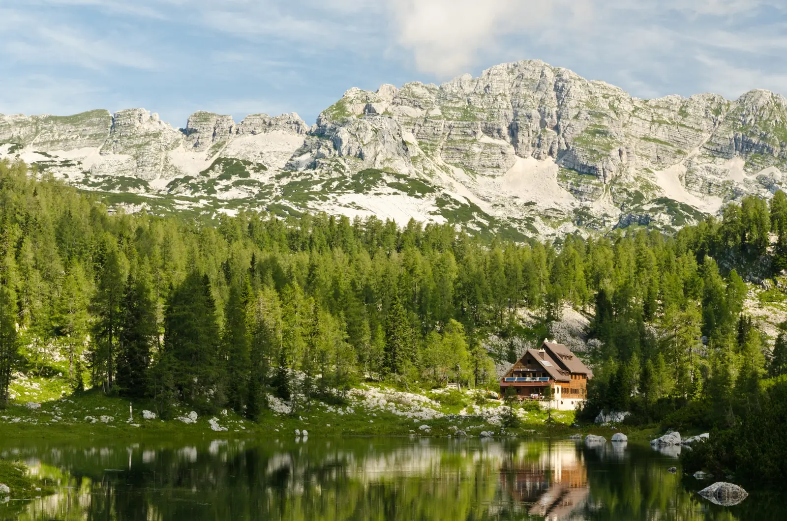 The Koča pri Triglavskih jezerih, a tall timber and white-walled mountain hut with a brown roof, standing among larch and spruce on the far shore of a still green lake that reflects it, with a long pale limestone ridge rising behind
