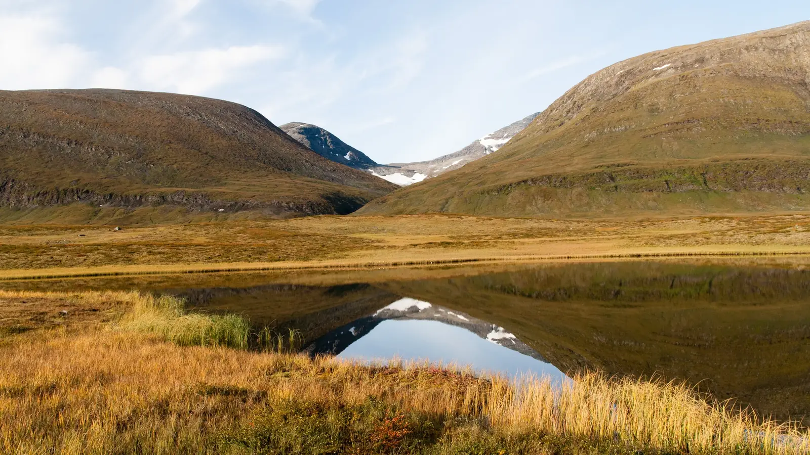 Autumn tundra in the Tjäktjavagge valley on the Kungsleden, with golden tussock grass and red dwarf shrubs in the foreground, a still pool reflecting the hills, and two rounded fells with small snowfields forming a wide notch beyond
