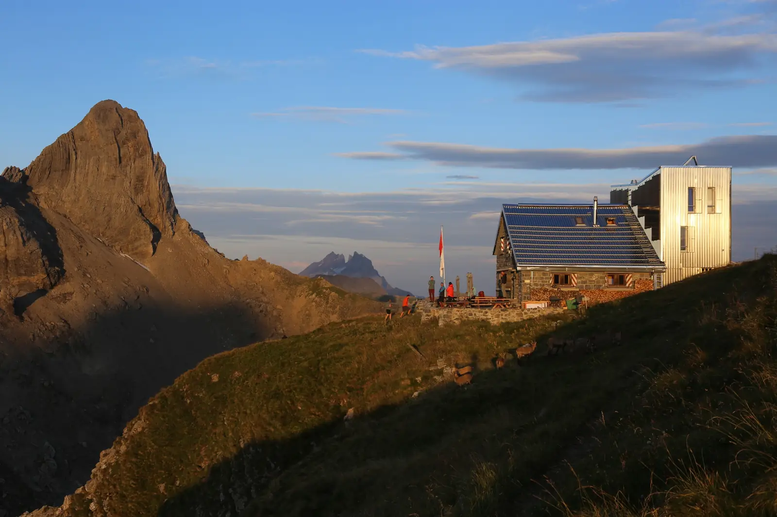 Cabane Rambert in low evening light — a stone hut with a solar-panelled roof beside a taller metal-clad annexe, walkers standing at picnic benches on its stone terrace beside a Swiss flag, wild goats grazing on the grassy slope below, and a sharp shadowed rock peak rising on the left