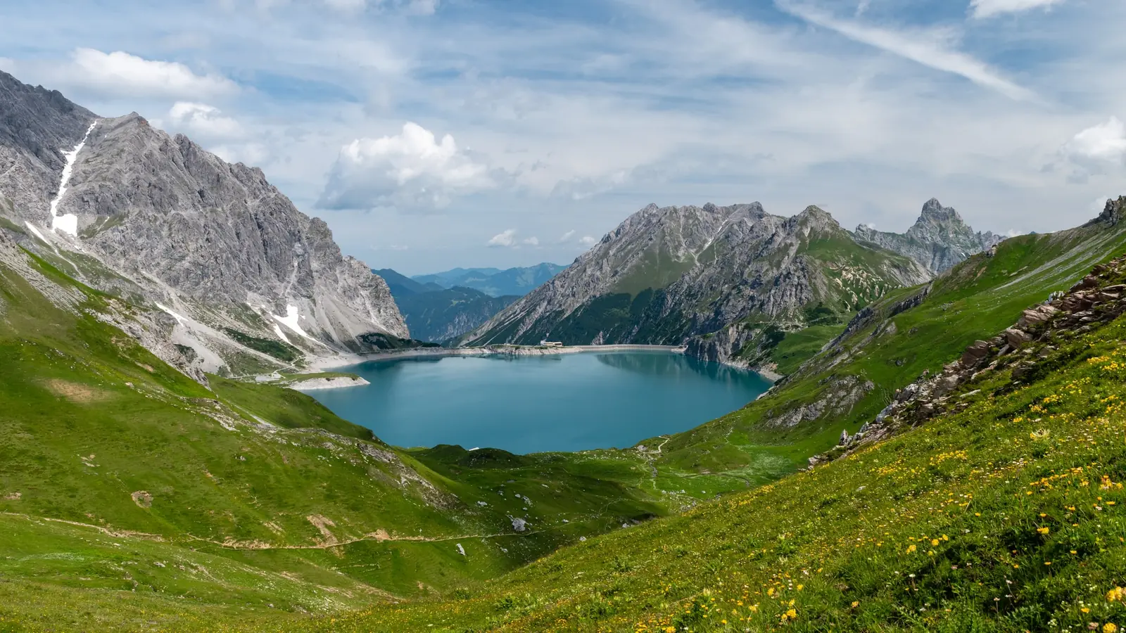 The turquoise Lünersee filling a limestone basin, seen from a flowering meadow above, with a curved pale dam wall and the small Douglasshütte at its far end and snow-streaked gray ridges rising on the left