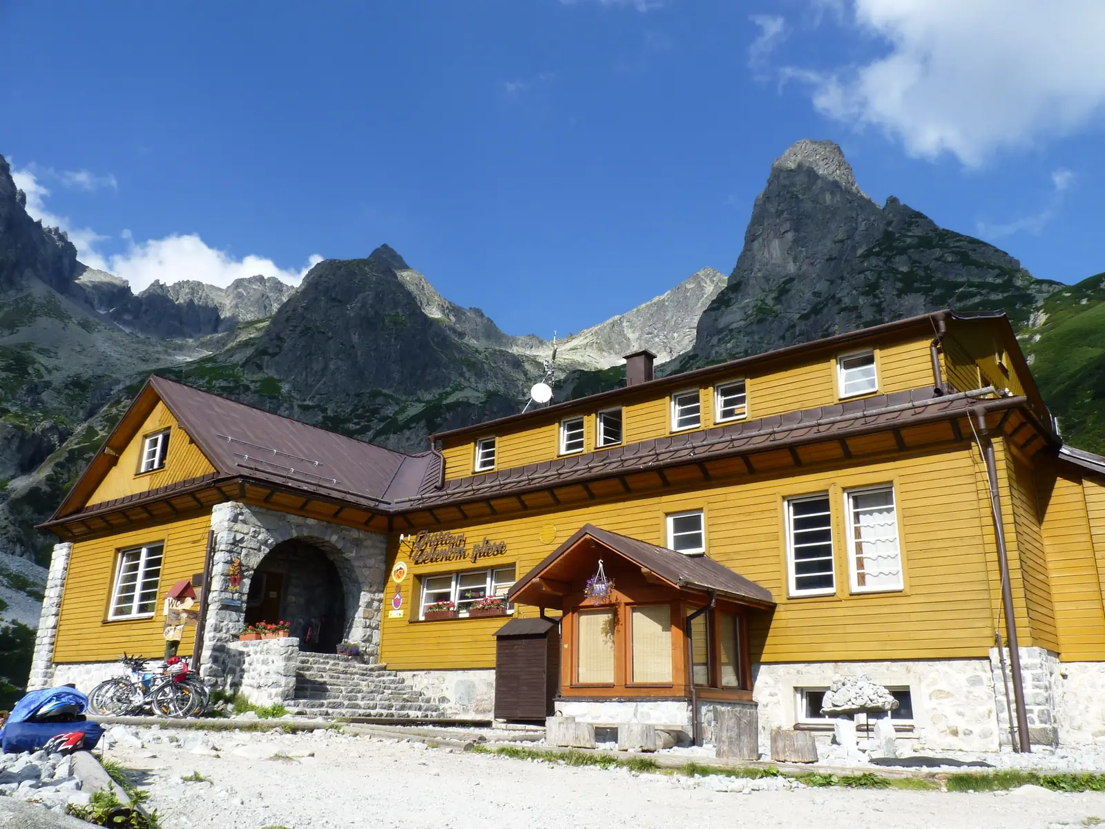 The Chata pri Zelenom plese, a long two-storey mountain hut clad in mustard-yellow timber over a gray stone base with a brown metal roof and red geraniums in the window boxes, with gray rock faces and a pointed rock tower rising behind it into a blue sky