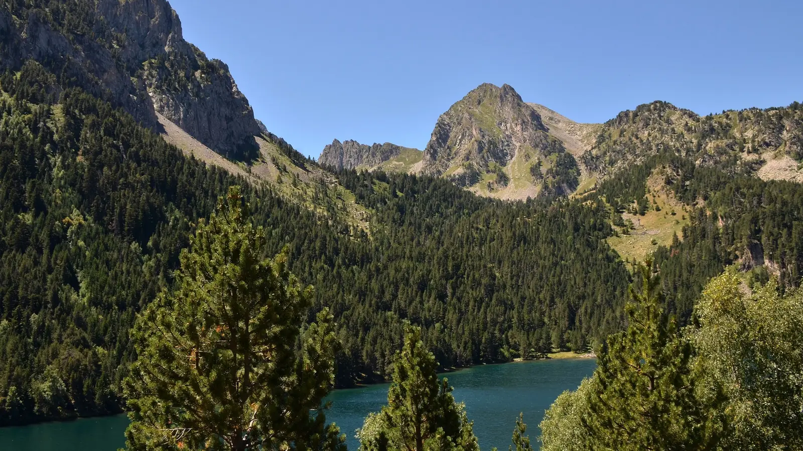 A turquoise lake below forested slopes and jagged granite peaks in the Aigüestortes i Estany de Sant Maurici national park in the Catalan Pyrenees
