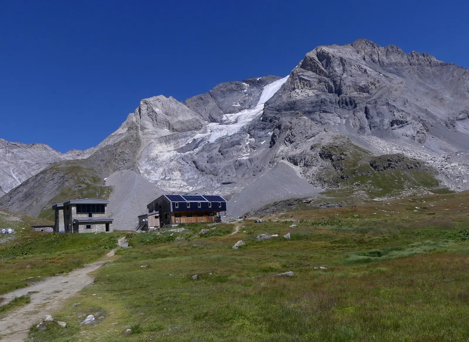 The modern timber and stone Refuge du Col de la Vanoise with solar panels on its roof, standing on grass below the gray rock face and hanging glacier of the Grande Casse