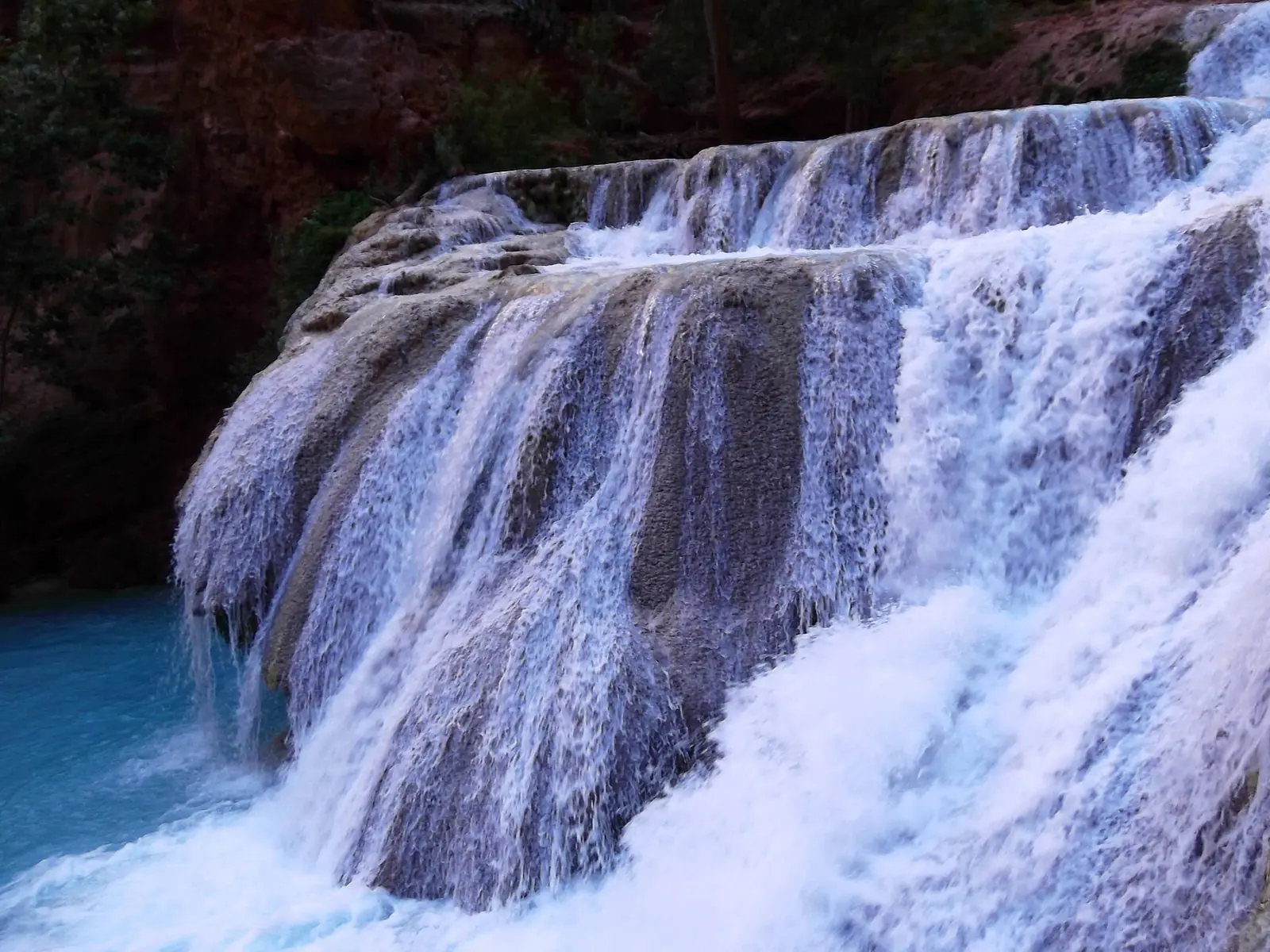 Beaver Falls spilling in wide shallow tiers over pale travertine ledges into a bright blue-green pool, dark canyon vegetation behind