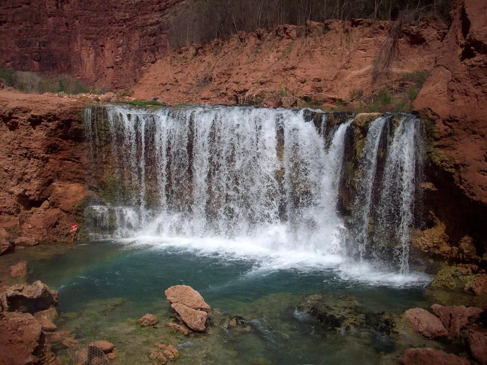 A wide curtain of white water spilling over a broad mossy travertine ledge into a green pool, framed by red canyon walls