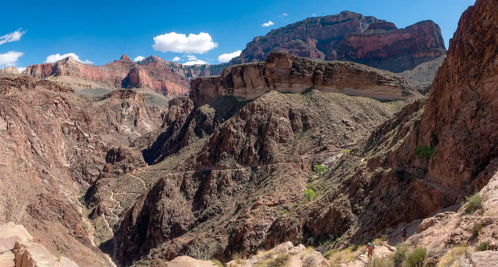 The Bright Angel Trail in Grand Canyon National Park threading in pale switchbacks across bare rock and scrub above Pipe Creek, with two tiny hikers on the trail at lower right and no shade anywhere in the frame