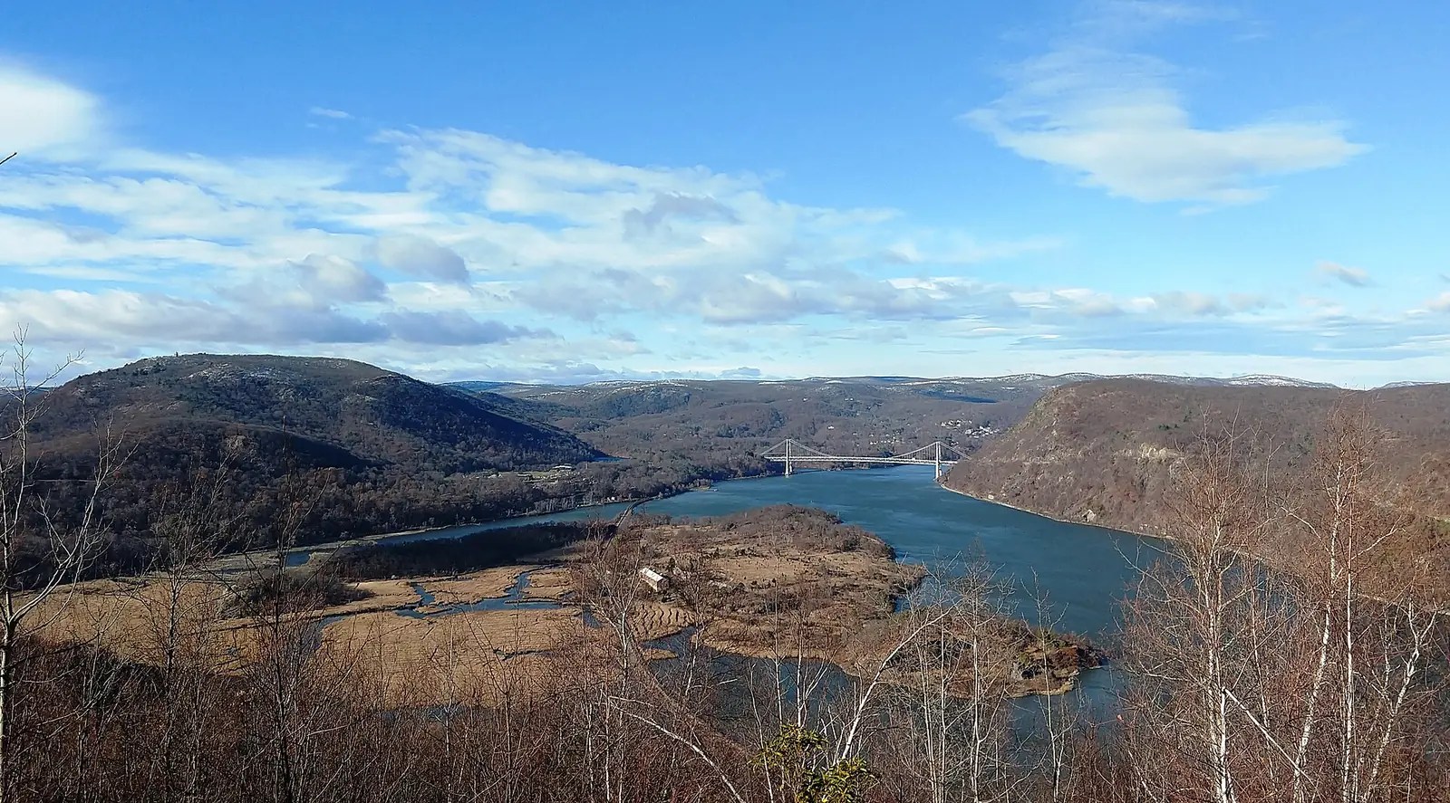 A high wide view over the Hudson River in late autumn with the Bear Mountain Bridge spanning it in the distance, bare brown ridges on both banks and a marshy flat in the foreground