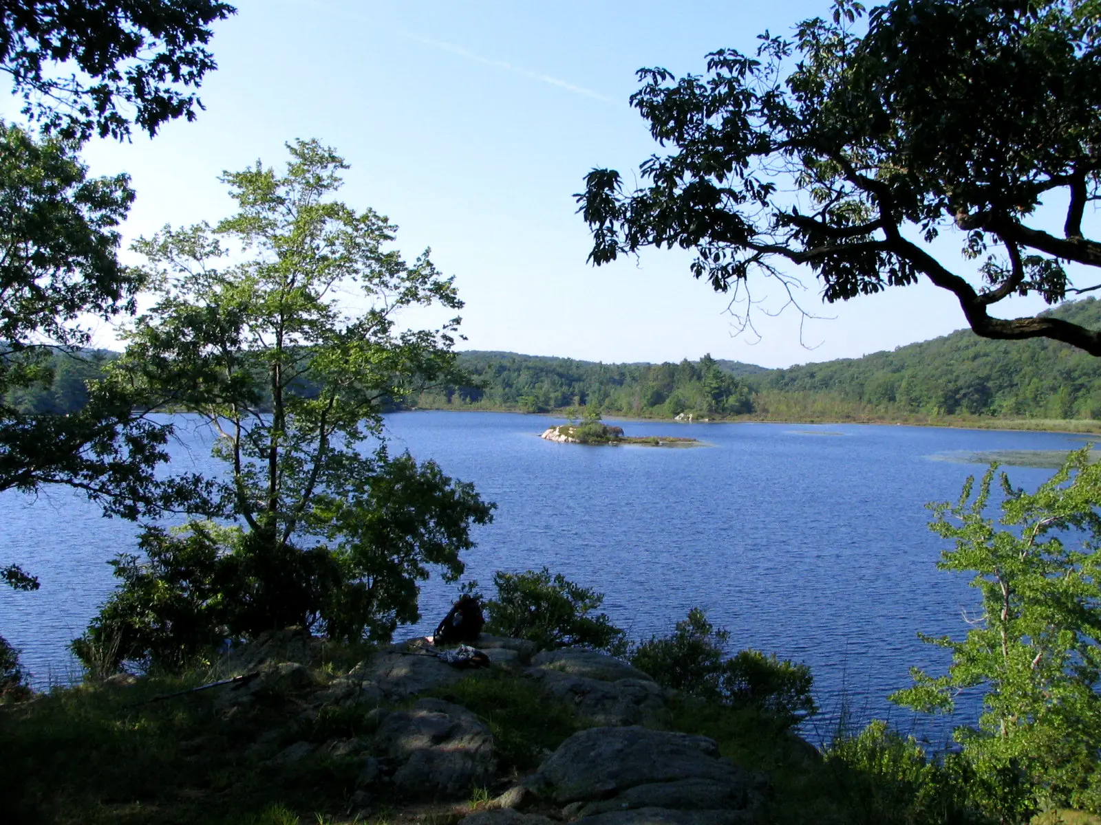 A blue pond ringed by low wooded hills seen from a rock ledge, with a hiker's dark pack resting on the rock in the foreground under a bright summer sky