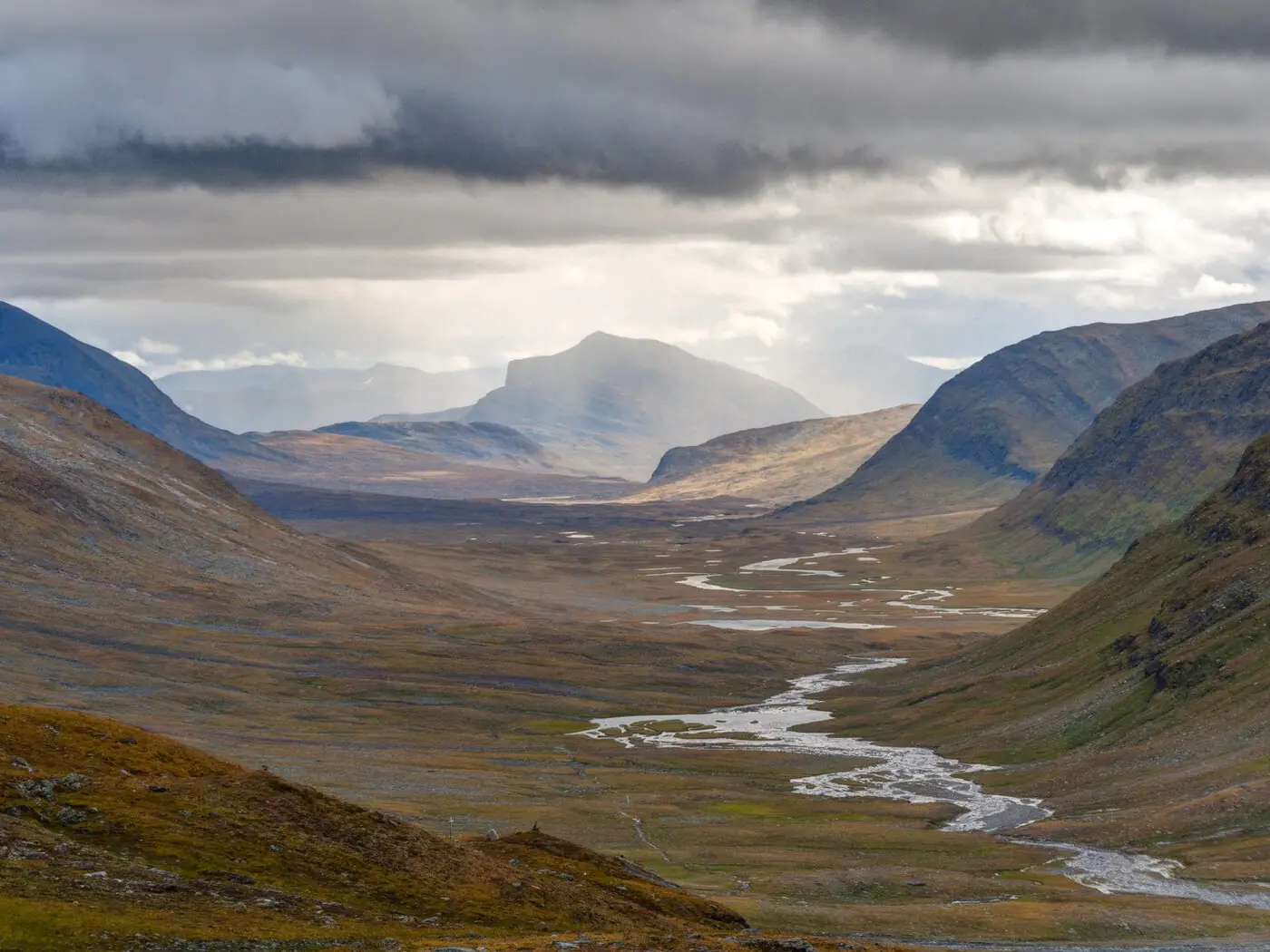 The wide U-shaped glacial valley of Tjäktjavagge in Swedish Lapland, a braided river threading its floor, autumn-coloured tundra on the slopes and storm light breaking through cloud