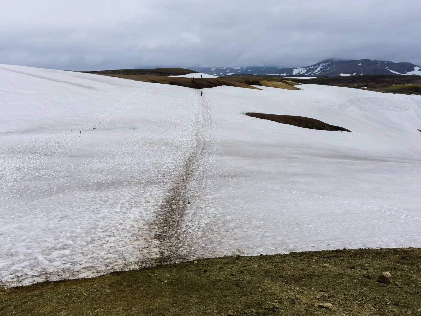 A broad snowfield on the Laugavegur trail with a single boot-worn track running across it, two tiny walkers in the distance and dark volcanic ridges under low gray cloud