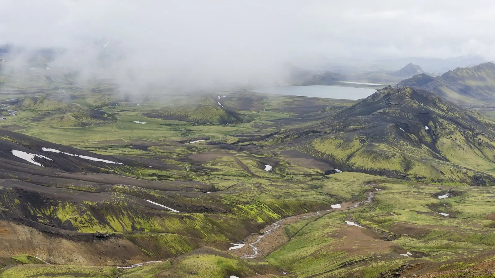 The Icelandic highlands from the Laugavegur trail, bright green moss over black volcanic ridges with a braided river and the pale lake of Álftavatn under low cloud