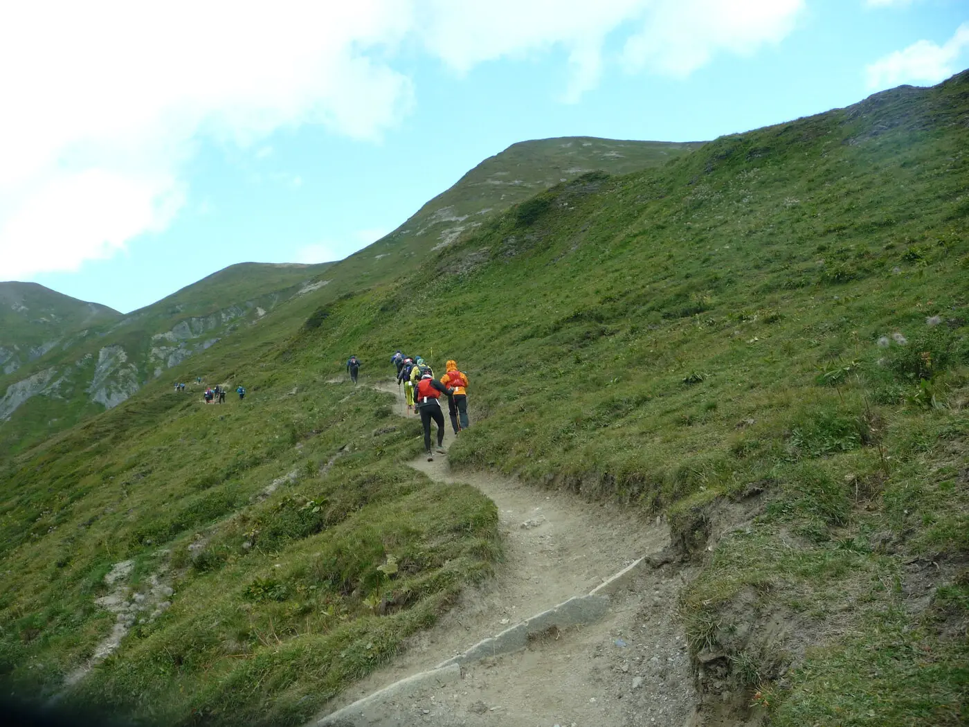 A line of runners in small vest packs strung out up a narrow dirt path on a grassy alpine slope, climbing towards the Grand Col Ferret during the 2021 Ultra-Trail du Mont-Blanc