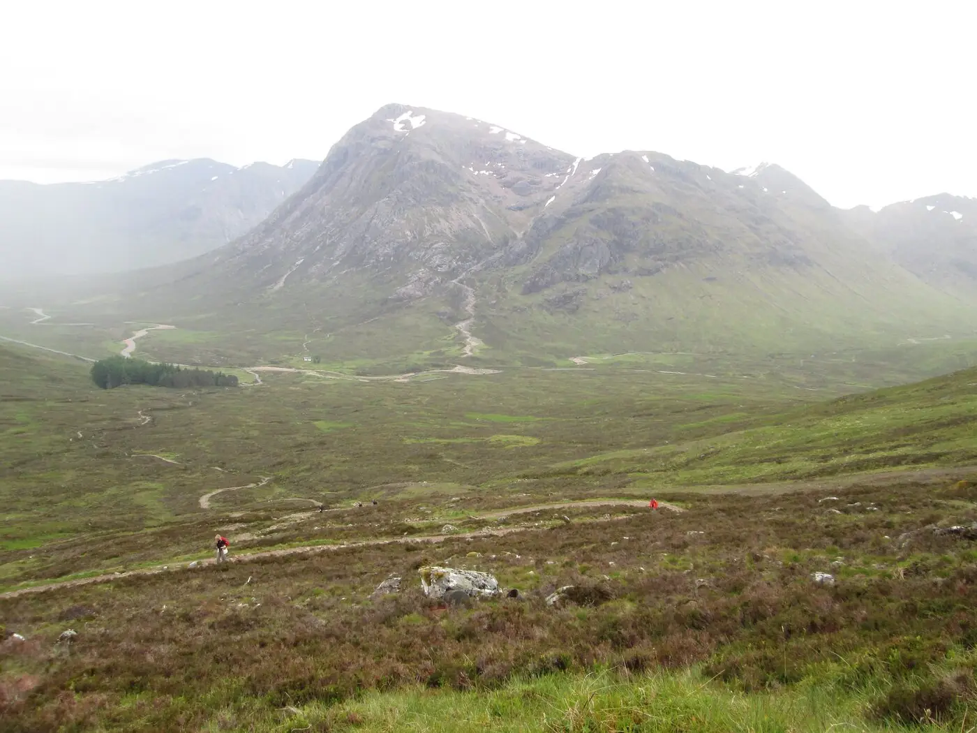The West Highland Way as a pale path across green and heather moorland near the Devil's Staircase, two small walkers on it, with the pyramid of Buachaille Etive Mor rising in hazy light beyond