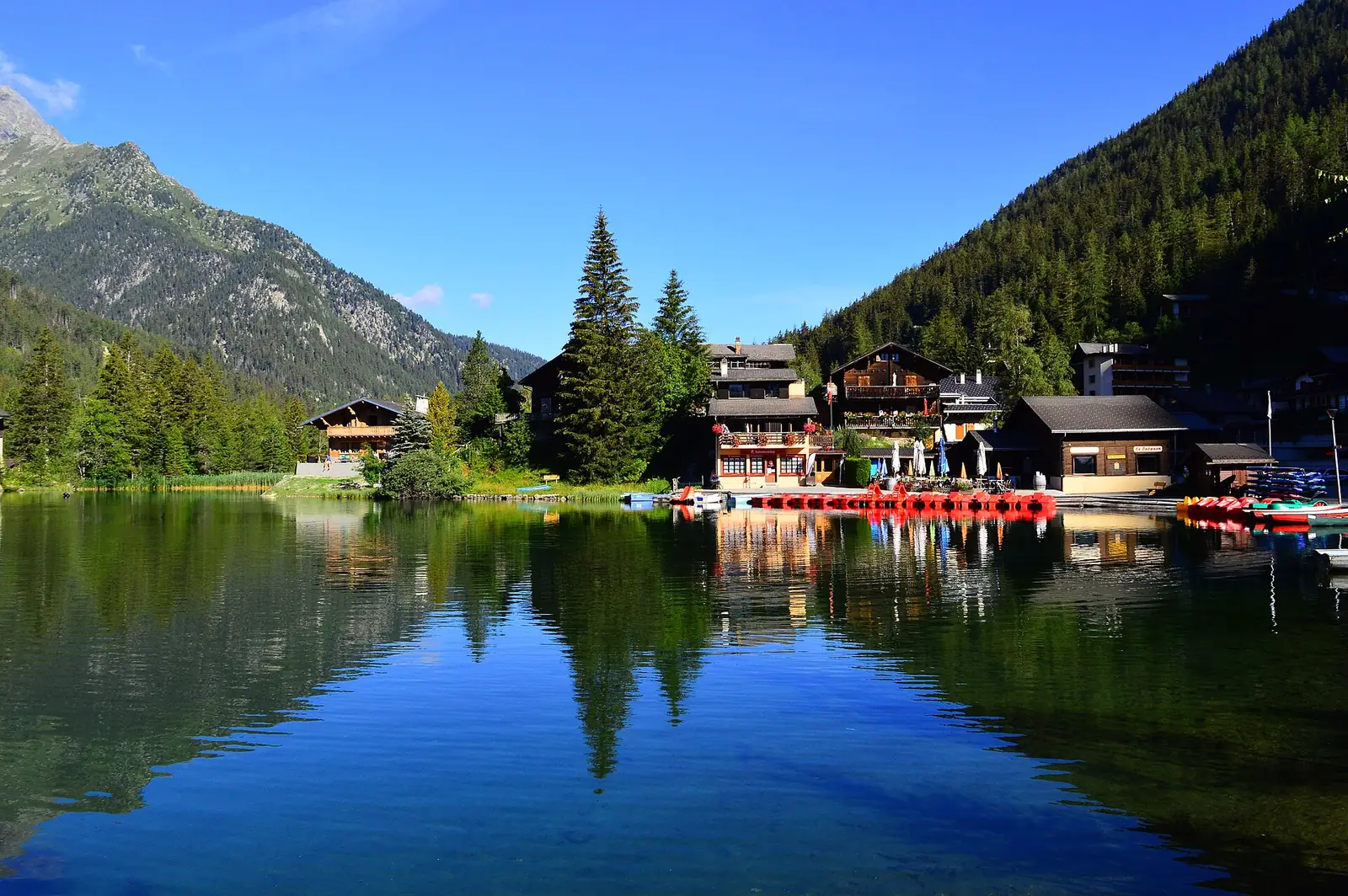 A still mountain lake reflecting wooden chalets, tall pine trees and a forested ridge under a deep blue sky