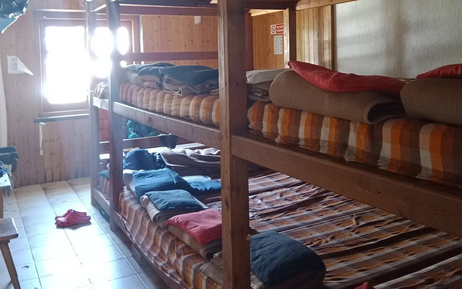 Wooden bunk platforms in a refuge dormitory with striped mattresses, folded blankets and pillows laid out at each place, and a pair of red hut slippers on the tiled floor
