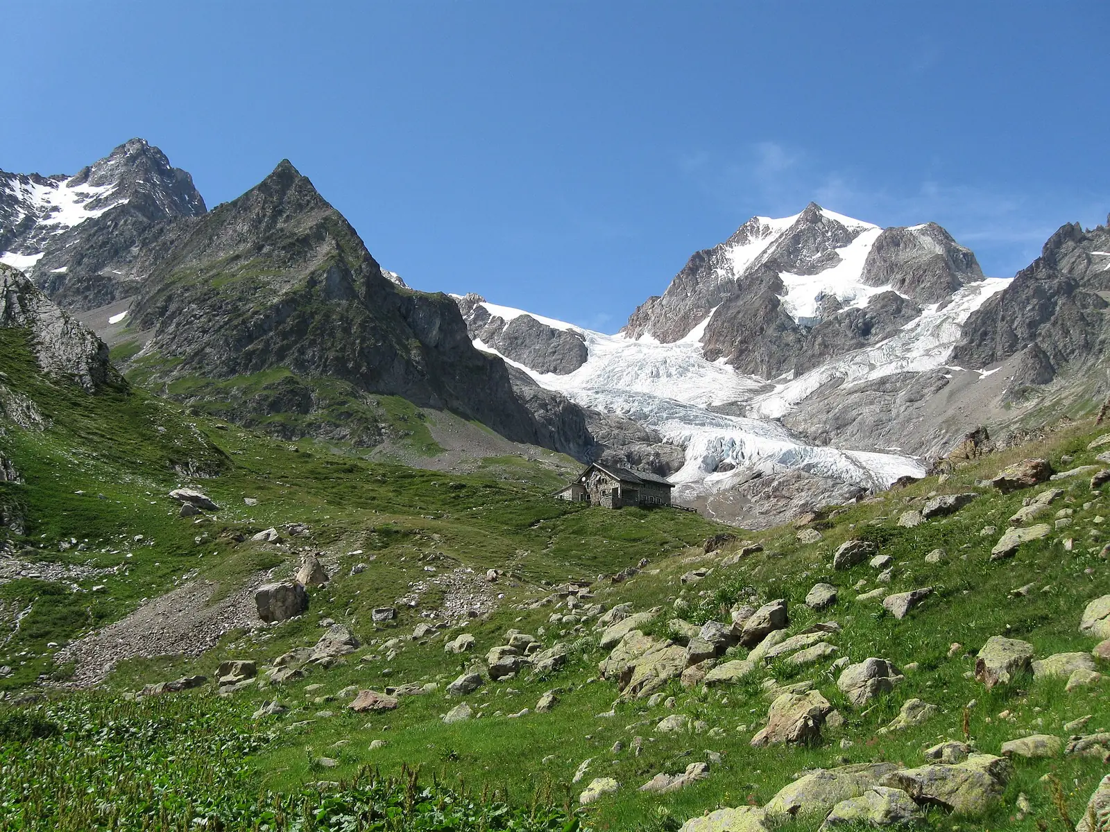 The small stone Rifugio Elisabetta sitting on a grassy, boulder-strewn slope below a broad tumbling glacier and dark rock peaks under a clear blue sky
