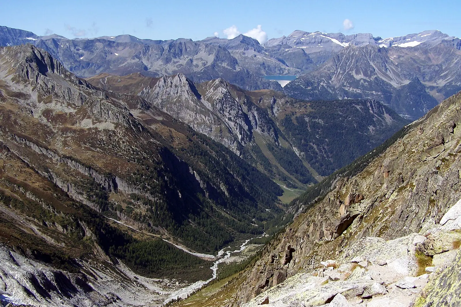 A view down from a high rocky pass over steep scree and cliffs into a deep valley, with layers of gray mountains and a small reservoir far in the distance