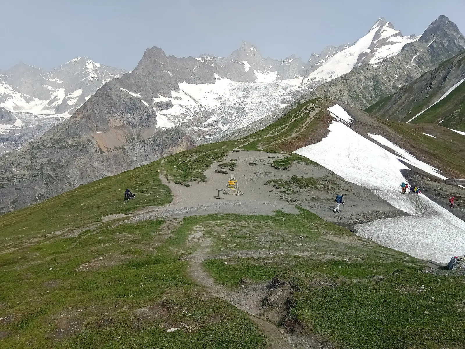 A grassy pass with a yellow trail signpost, a few hikers crossing a lingering snowfield on the right, and glaciated peaks behind