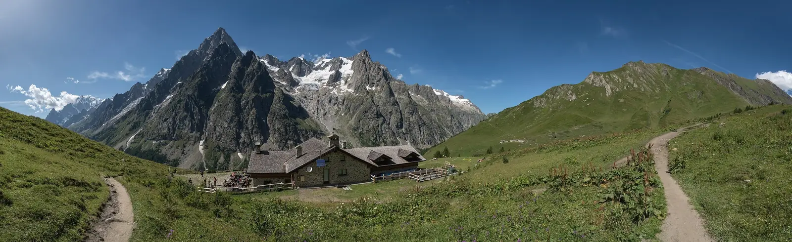 The stone Rifugio Bonatti on a green alpine shoulder in Italy's Val Ferret, with a dirt trail curving past it and the granite spires and glaciers of the Grandes Jorasses rising behind