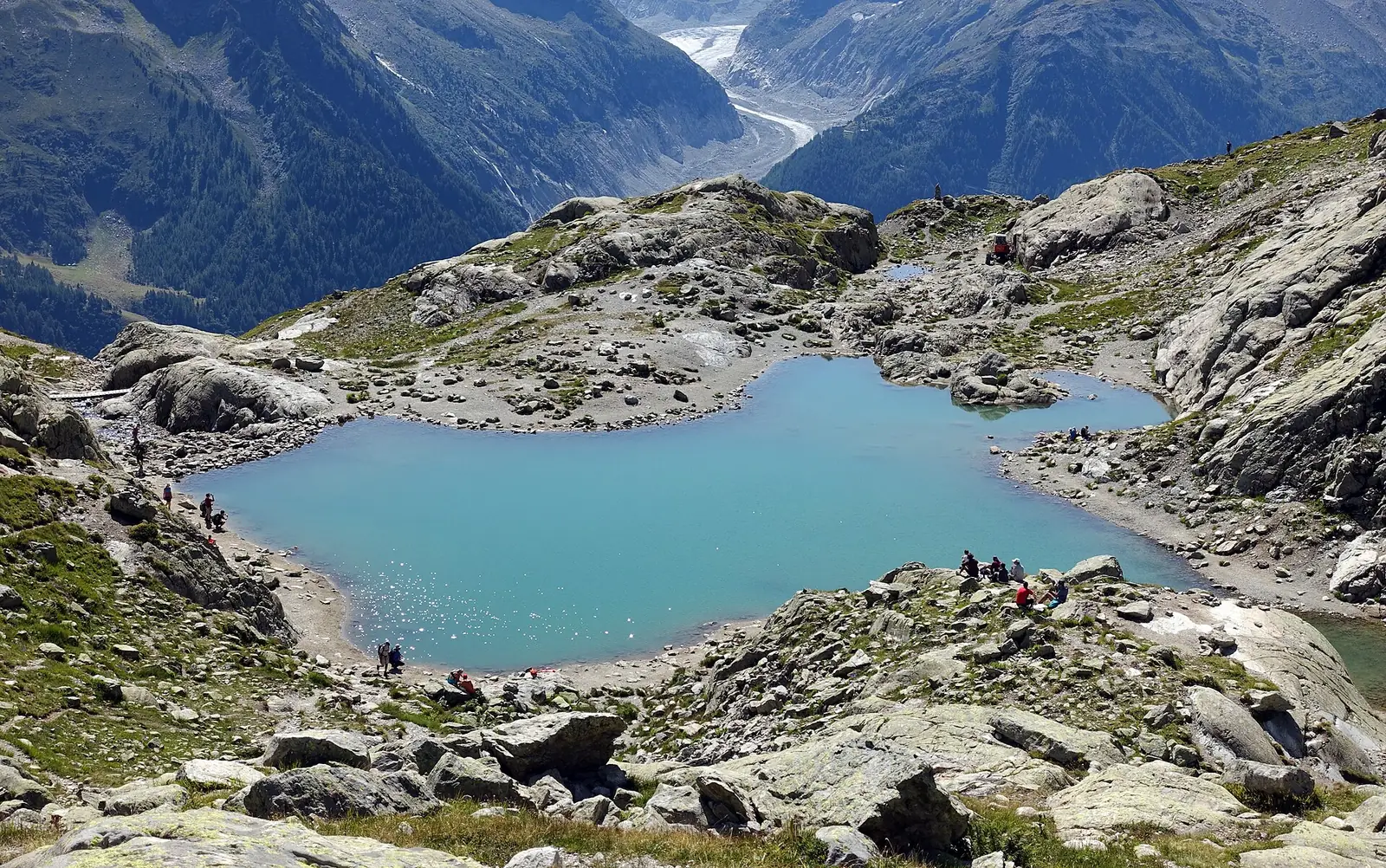 A turquoise alpine lake ringed by gray granite slabs, with small groups of hikers resting on the rocks and a glacier filling the valley far below